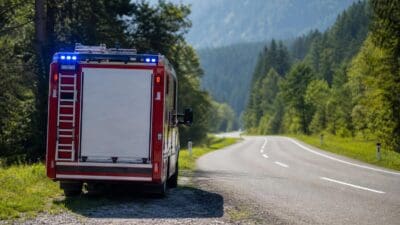 Heckansicht eines roten Feuerwehrfahrzeugs mit aktiviertem Blaulicht am Straßenrand in bergiger Landschaft