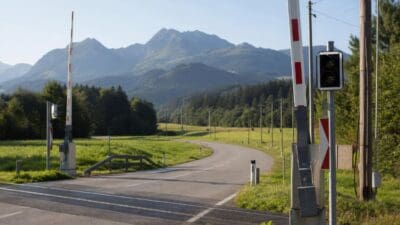 Rot-weiße Schranken eines Bahnübergangs an einer kurvigen Landstraße vor bewaldeten Bergen