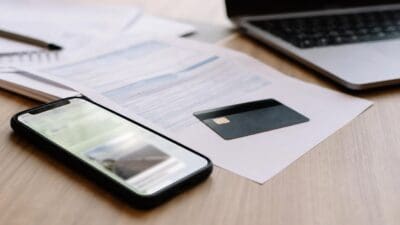 Smartphone liegt auf Dokumenten neben schwarzer Kreditkarte auf Holztisch mit Laptop