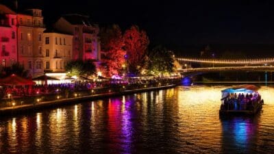 Beleuchtetes Boot auf der Drau in Villach bei Nacht mit Menschen am Ufer und bunten Lichtern