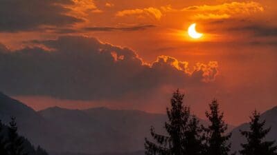 Partielle Sonnenfinsternis in orangem Himmel über Bergkette mit Tannen im Vordergrund