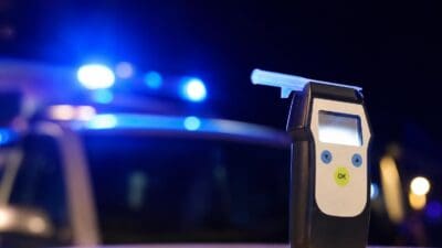 Nahaufnahme eines digitalen Atemalkoholmessgeräts mit blau beleuchtetem Mundstück vor verschwommenem Blaulicht eines Polizeifahrzeugs