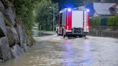 Rotes Feuerwehrauto mit Blaulicht fährt durch überflutete Straße neben Steinmauer