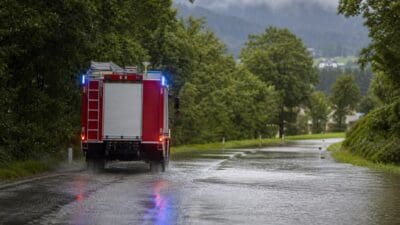 Rotes Feuerwehrauto mit Blaulicht fährt auf überfluteter Straße in grüner Berglandschaft
