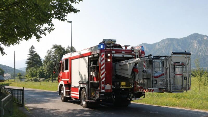 Rotes und weißes Feuerwehrfahrzeug mit ausgefahrener Arbeitsbühne auf einer Landstraße vor Bergen