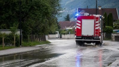 Heckansicht eines roten Feuerwehrfahrzeugs mit Blaulicht auf überfluteter Straße vor Häusern und Bergen