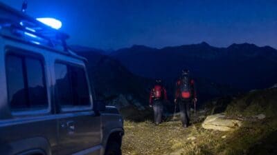 Zwei Bergretter mit Rucksäcken und Stöcken gehen neben einem Fahrzeug mit Blaulicht in den Bergen