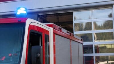 Rotes Feuerwehrfahrzeug mit aktiviertem blauem Blaulicht vor einer Feuerwache mit Glasfront