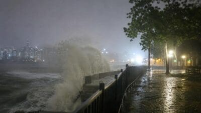 Hohe Wellen brechen bei Nacht über ein Geländer auf einer nassen Uferpromenade mit Stadtlichtern im Hintergrund.