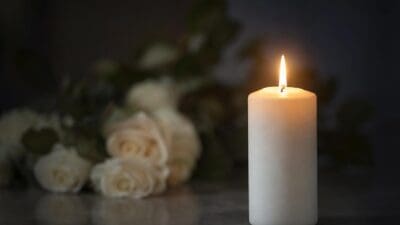 Eine brennende weiße Kerze vor einem unscharfen Strauß weißer Rosen.