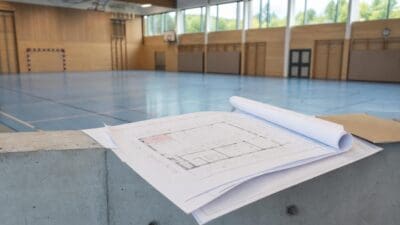 Gerollte und ausgebreitete Baupläne liegen auf einer Betonmauer vor einer leeren Sporthalle.
