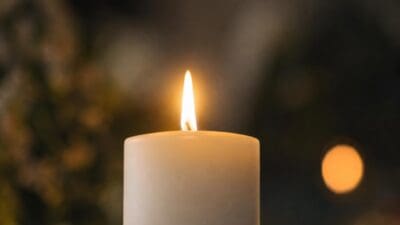 Eine brennende weiße Kerze mit heller Flamme vor unscharfem Hintergrund.