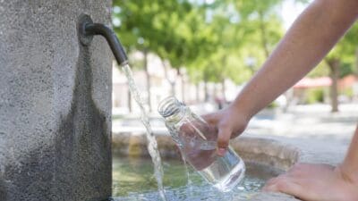 Eine Hand hält eine Glasflasche unter fließendes Wasser aus einem Hahn an einer Steinmauer.
