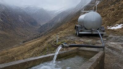Wasser fließt aus einem Schlauch eines silbernen Tankwagens in einen steinernen Trog in den Bergen.
