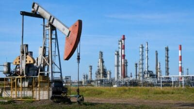 Eine Ölpumpe mit rotem Gegengewicht steht vor einer Raffinerie mit vielen Schornsteinen