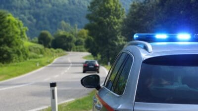 Silbernes Polizeifahrzeug mit blauem Blaulicht auf kurviger Landstraße in grüner Hügellandschaft mit weiterem Auto im Hintergrund