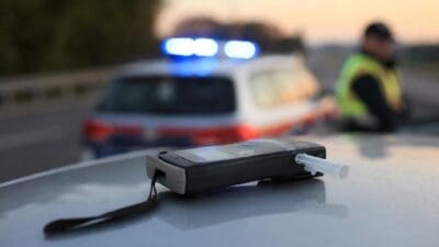 Nahaufnahme eines Alkoholtesters auf einer Autohaube mit unscharfem Polizeiwagen und Blaulicht im Hintergrund.