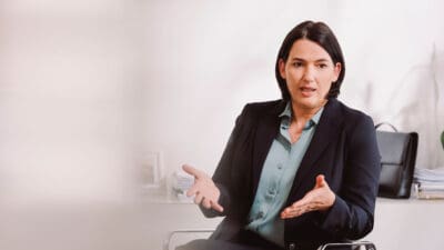 Dunkelhaarige Frau in dunklem Blazer gestikuliert mit beiden Händen am Schreibtisch.