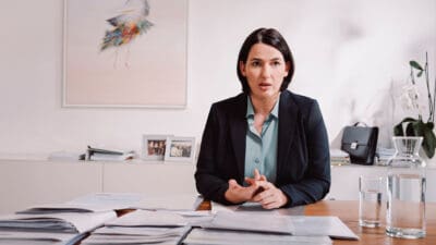Eine Frau mit dunklen Haaren in einem schwarzen Blazer sitzt an einem Schreibtisch mit Papieren.
