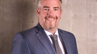 Lächelnder Mann mit grauem Haar und Bart in blauem Anzug mit Krawatte vor Betonwand