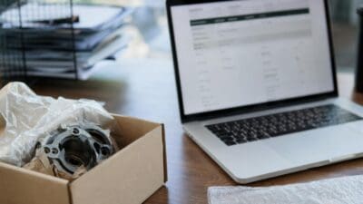 Metallisches Fahrzeugteil in einem offenen Karton neben einem Laptop auf einem Holztisch