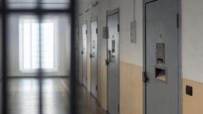 Langer Flur in einer Justizanstalt mit mehreren grauen Metalltüren und Gitterstäben im Vordergrund