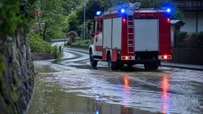 Rotes Feuerwehrfahrzeug mit Blaulicht fährt durch überflutete Straße bei Dämmerung