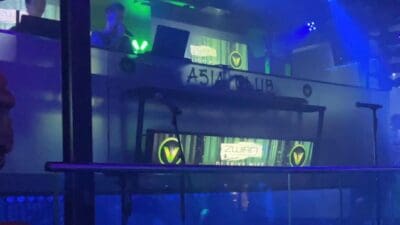 DJ-Pult in einem Nachtclub mit blauen und grünen Scheinwerfern, V-Logos auf Bildschirmen und Bühne