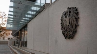 Metallener Bundesadler mit ausgebreiteten Schwingen an einer hellen Betonwand neben Glasüberdachung.