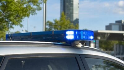 Blaues Blaulicht auf dem Dach eines silbernen Polizeifahrzeugs vor Bäumen und Gebäuden