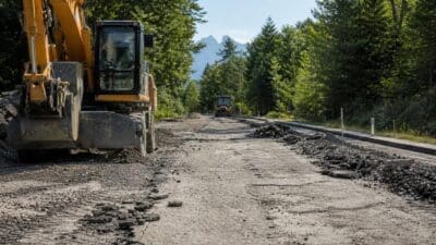 Großer gelber Bagger auf einer aufgerissenen Landstraße bei Bauarbeiten mit Bergen im Hintergrund.