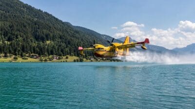 Ein gelbes Löschflugzeug fliegt über einem türkisfarbenen Bergsee und nimmt Wasser auf.