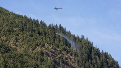 Hubschrauber mit Löschbehälter fliegt über bewaldetem Berg und löscht Waldbrand mit Wasser