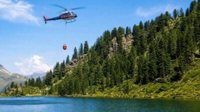 Ein Hubschrauber mit einem roten Löschbehälter am Seil fliegt über einem Bergsee.