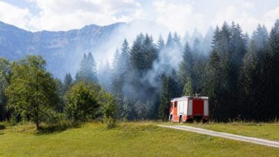 Rotes Feuerwehrauto steht auf einer Wiese vor einem rauchenden Wald mit Bergen im Hintergrund.