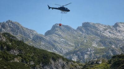 Hubschrauber mit rotem Löschbehälter fliegt über dem Karwendelgebirge bei Reith bei Seefeld