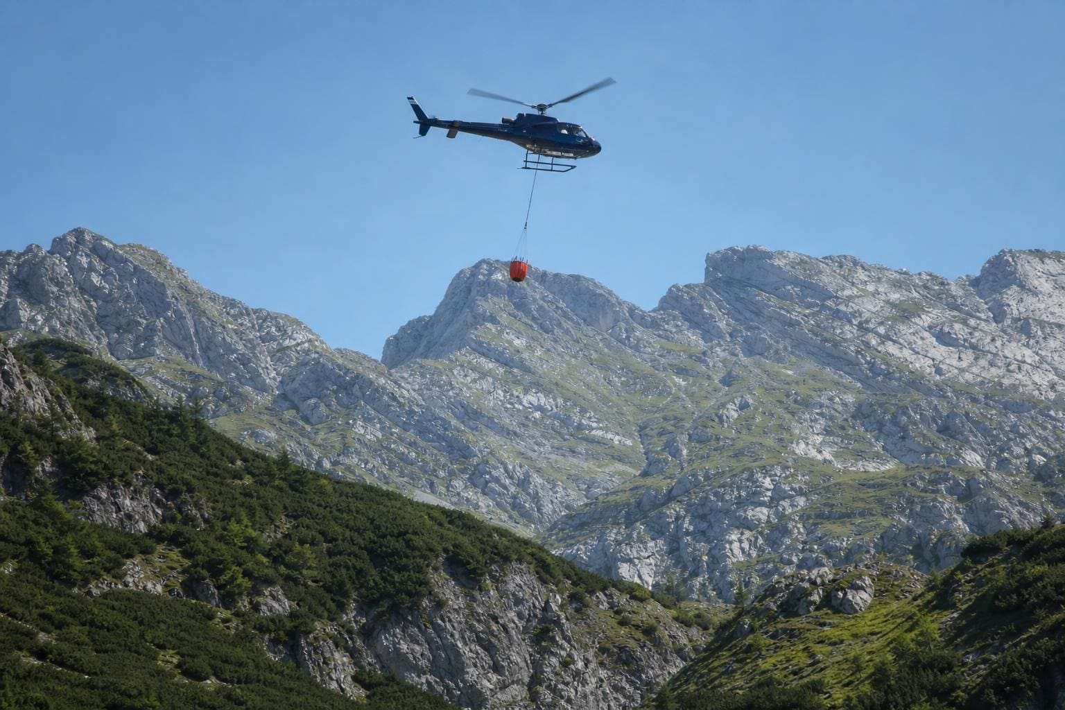 Hubschrauber mit rotem Löschbehälter fliegt über dem Karwendelgebirge bei Reith bei Seefeld
