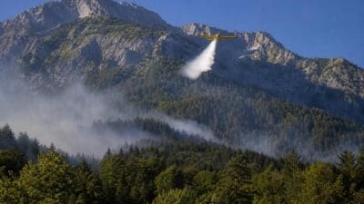 Gelbes Löschflugzeug fliegt über bewaldeten Bergen und versprüht weißes Löschmittel bei Srednji Vrh