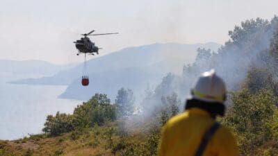 Feuerwehrmann in gelber Schutzkleidung beobachtet Löschhubschrauber mit Wasserbehälter über rauchigem Wald am Gardasee