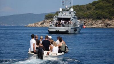 Menschen in einem kleinen Motorboot auf dem Meer vor einer felsigen Küste mit einem Schiff im Hintergrund