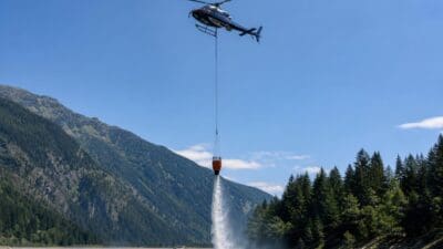 Ein Hubschrauber mit orangefarbenem Löschwasserbehälter nimmt Wasser aus einem Bergsee auf.