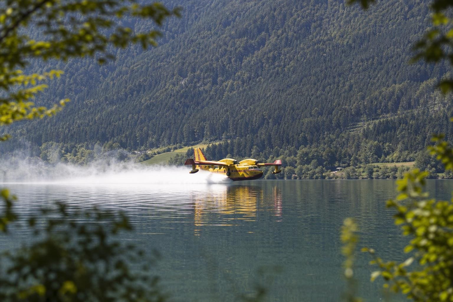 Gelbes Löschflugzeug gleitet über Bergsee und erzeugt große weiße Wassersprühwolke vor bewaldeten Bergen