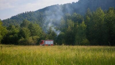 Rotes Feuerwehrfahrzeug auf einer Wiese vor bewaldetem Berg mit aufsteigendem Rauch