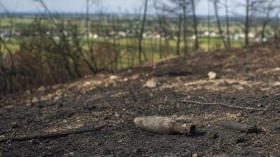 Rostiges Munitionsteil liegt auf verkohltem Boden zwischen verkohlten Ästen