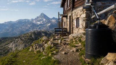 Alpenhütte aus Holz und Stein mit schwarzem Wassertank vor schneebedeckten Bergen