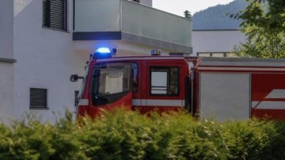 Rotes Löschfahrzeug mit eingeschaltetem Blaulicht steht hinter grünen Büschen vor einem Haus