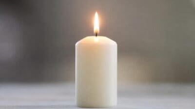 Eine brennende weiße Kerze mit heller Flamme auf einer hellen Oberfläche vor unscharfem Hintergrund.