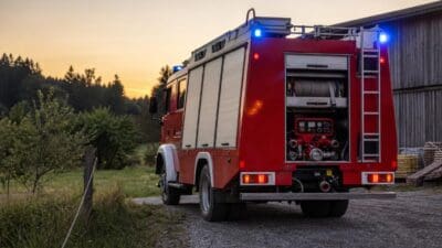 Heck eines roten Feuerwehrfahrzeugs mit offenen Abteilen und blauen Blinklichtern auf einem Feldweg.