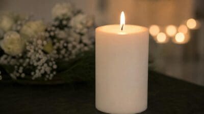 Eine brennende weiße Kerze auf einer dunklen Oberfläche mit unscharfen weißen Blumen im Hintergrund.