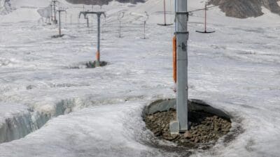 Grauer Metallmast eines Skilifts mit freigelegtem felsigem Sockel auf schmelzendem Gletschereis und weiteren Masten im Hintergrund